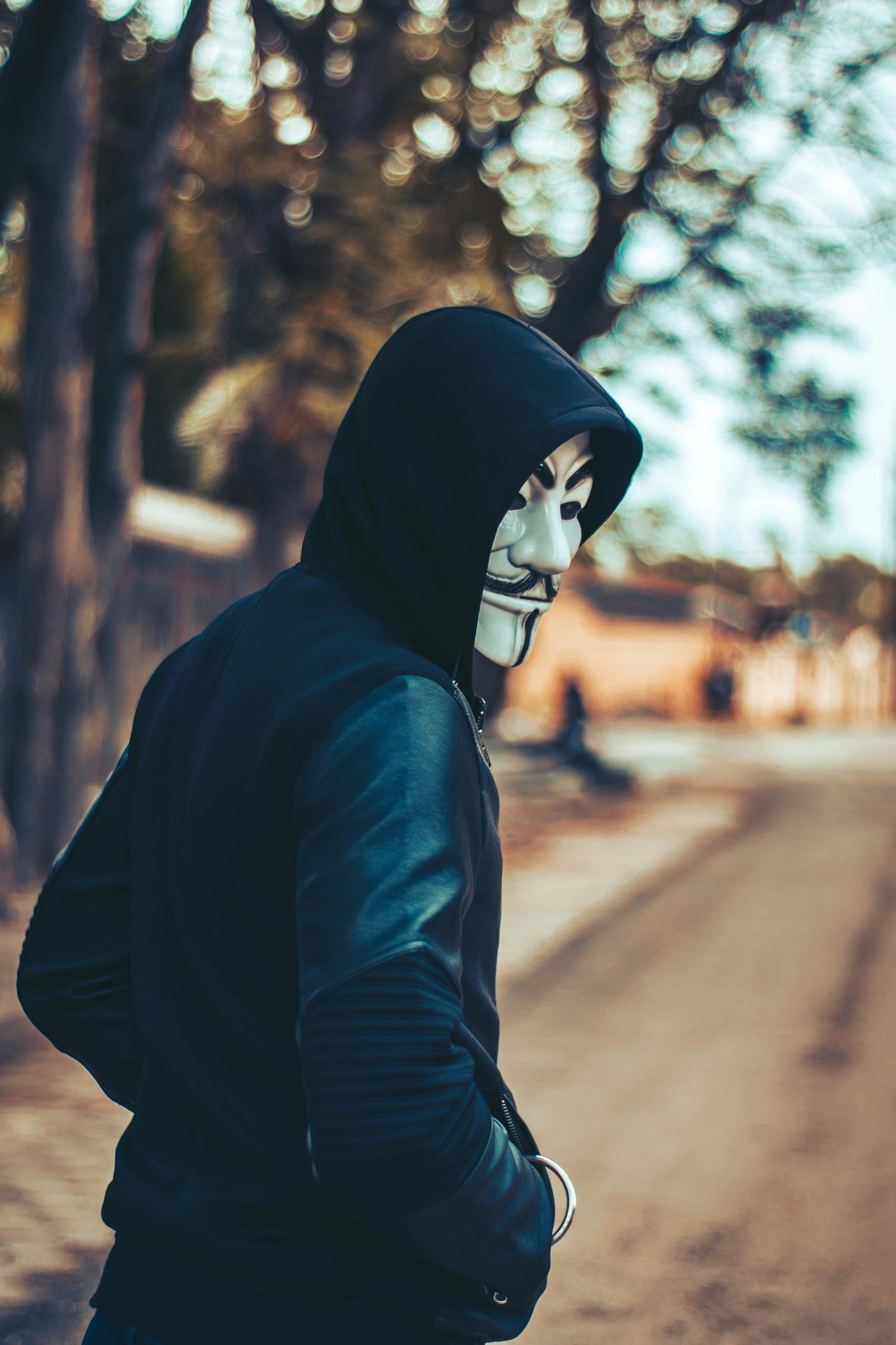 A man in a hoodie wearing a Guy Fawkes mask walks down an urban street, creating an air of mystery and intrigue.