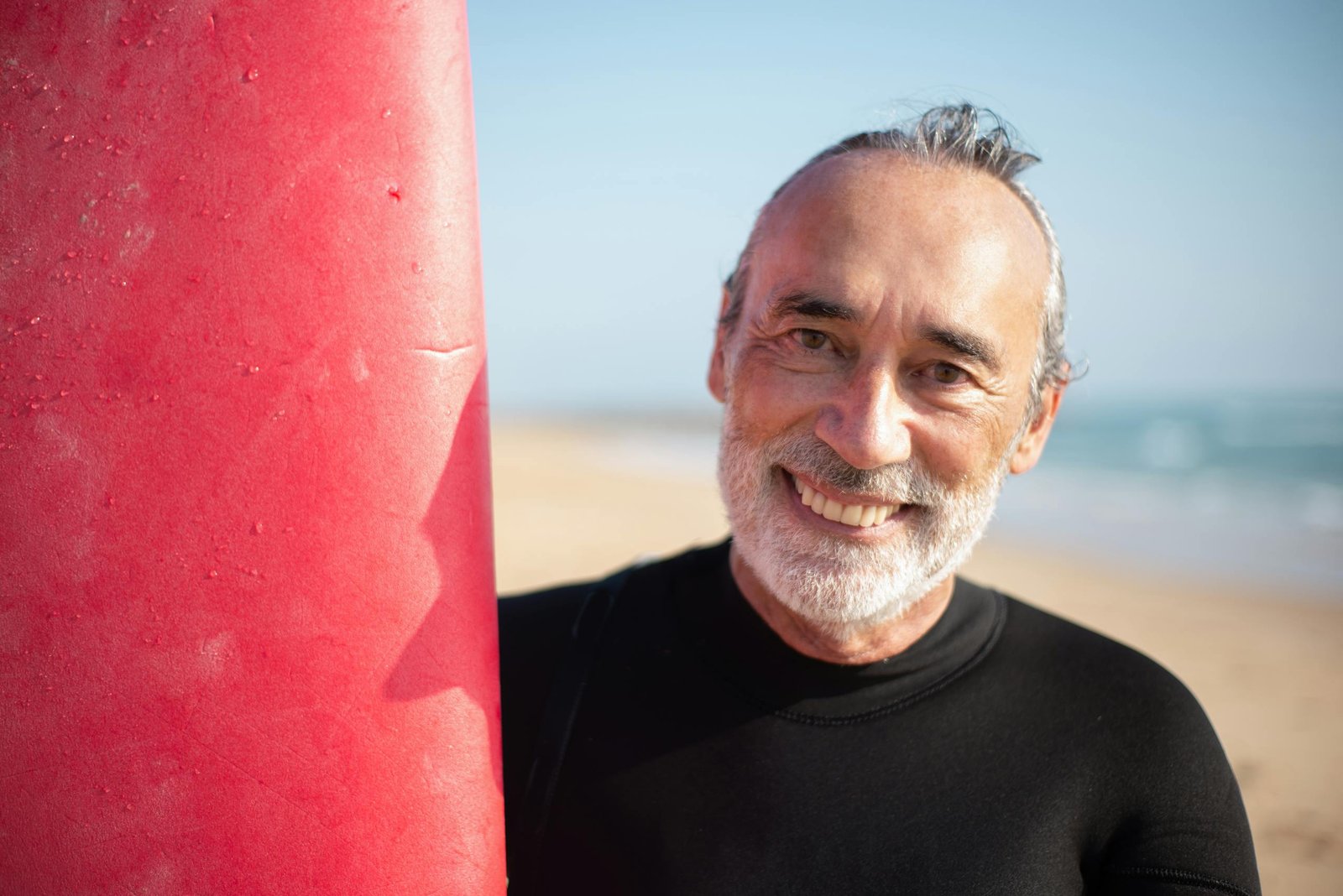 Smiling man at the beach holding a red surfboard—active, healthy lifestyle.