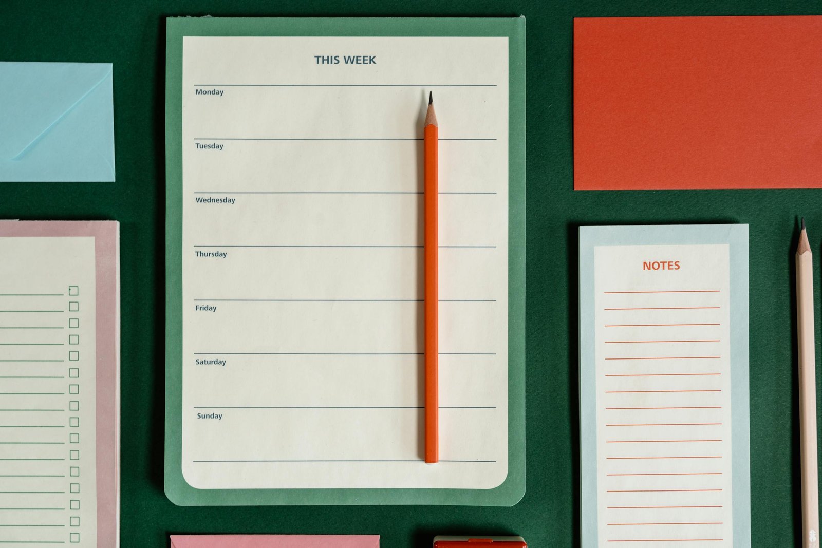 Flat lay of weekly planner, notes, envelopes, and pencils on a green background representing organization.