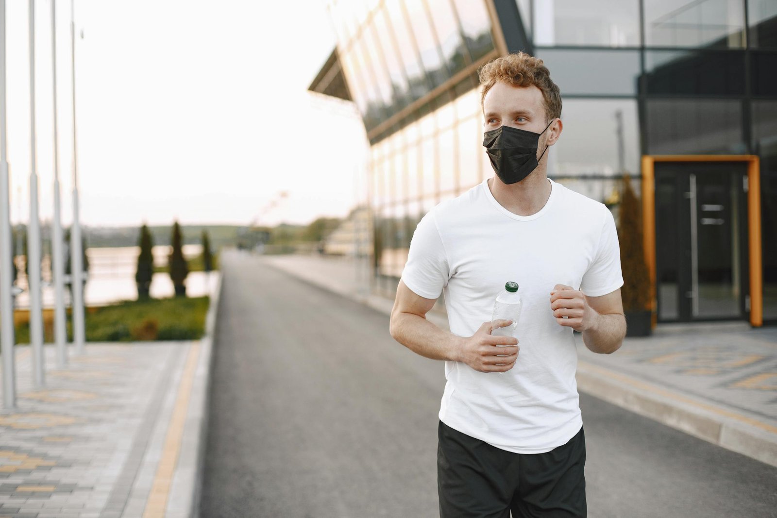 Man wearing a mask jogging with a water bottle on a modern urban road.