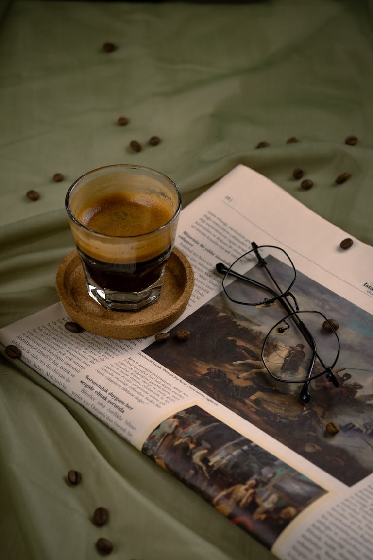 Glass of espresso on saucer, newspaper, and eyeglasses elegantly arranged.